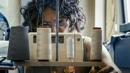 An older woman works intently at a sewing machine glimpsed between the many spools of thread