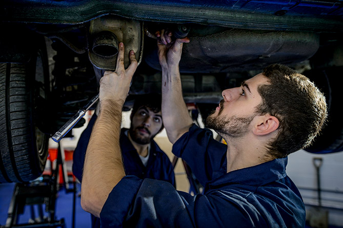 A bearded man stands under an elevated car servicing the exhaust while in the background his colleague holds a light